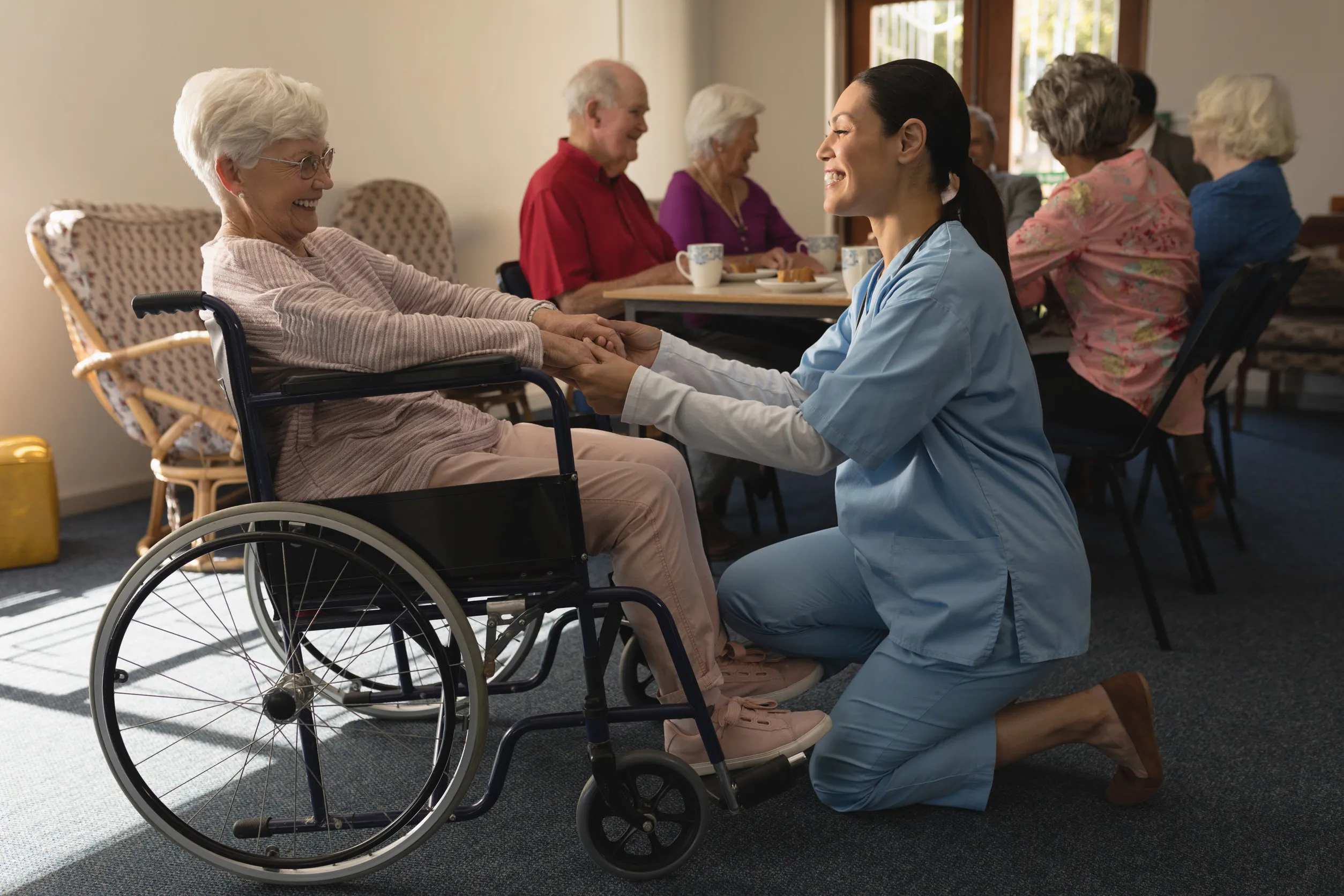 Side view of happy female doctor holding hands and talking with disable senior woman at home